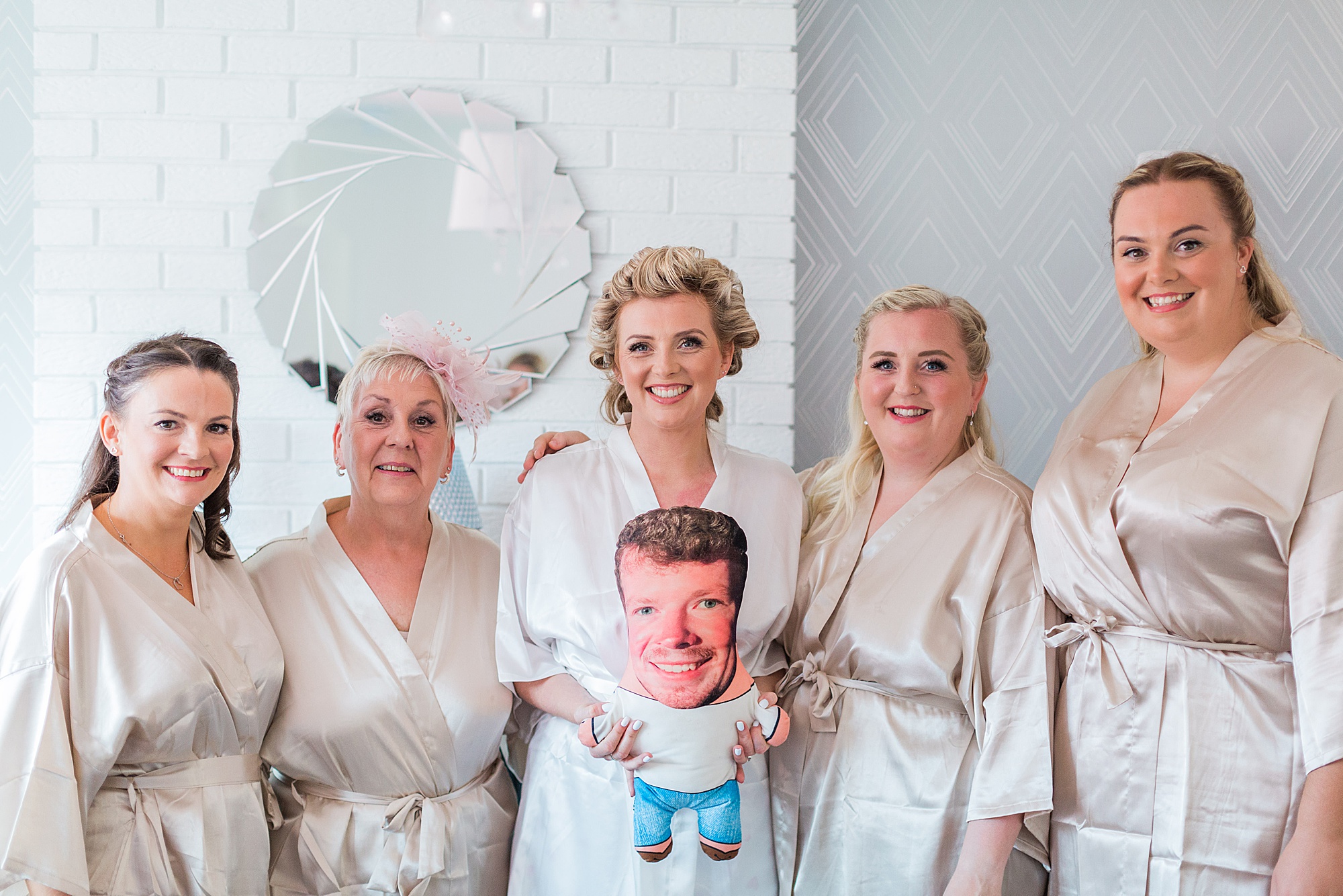 bride and bridesmaids in matching dressing gowns on the morning of the wedding holding a photo cushion of a husband who couldn't be at the wedding. All girls smiling with hair and makeup preparations underway 
