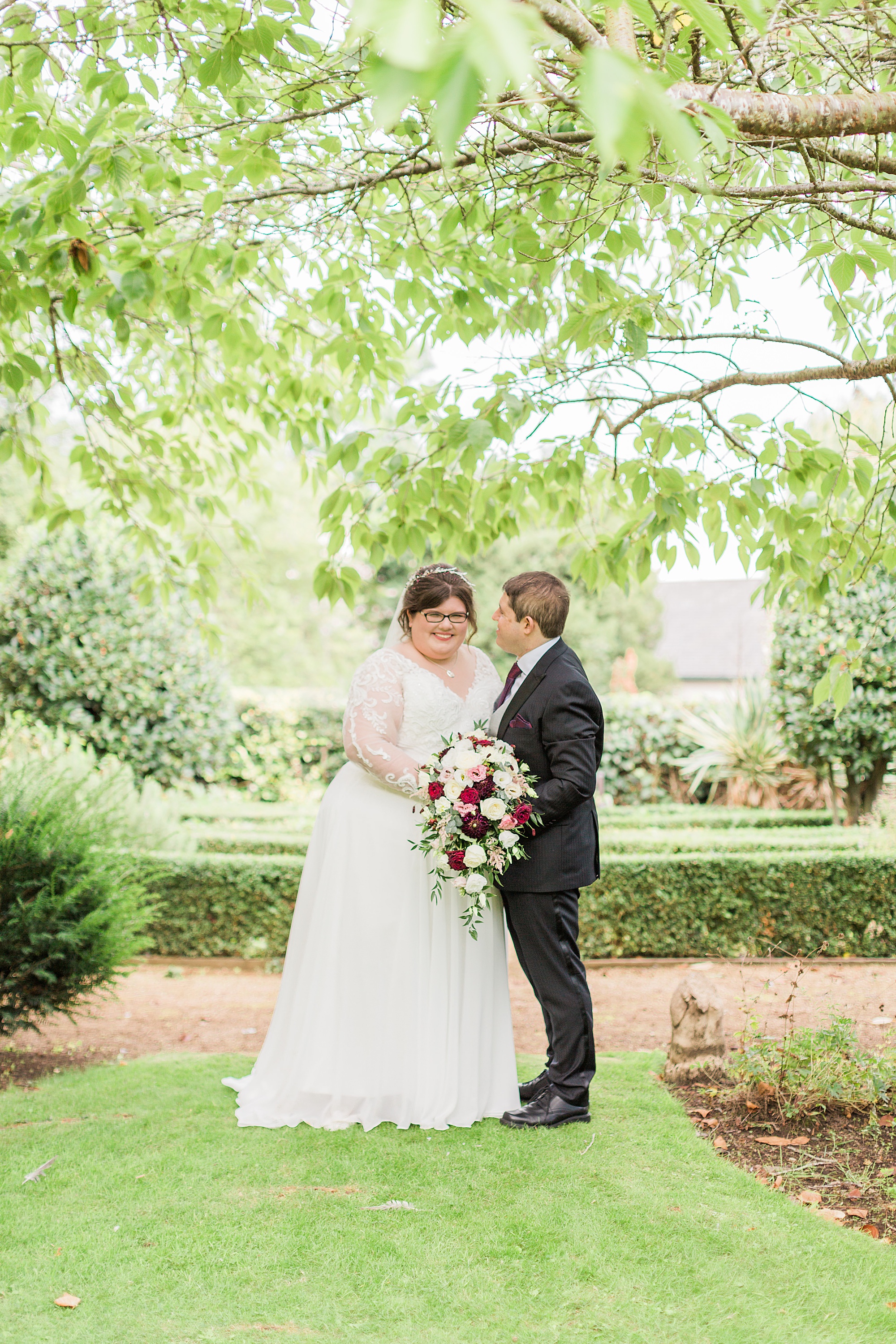 photo of a bride and groom at their wedding surrounded by trees and greenery, groom is facing in towards his bride and they're joined hands by her bouquet 