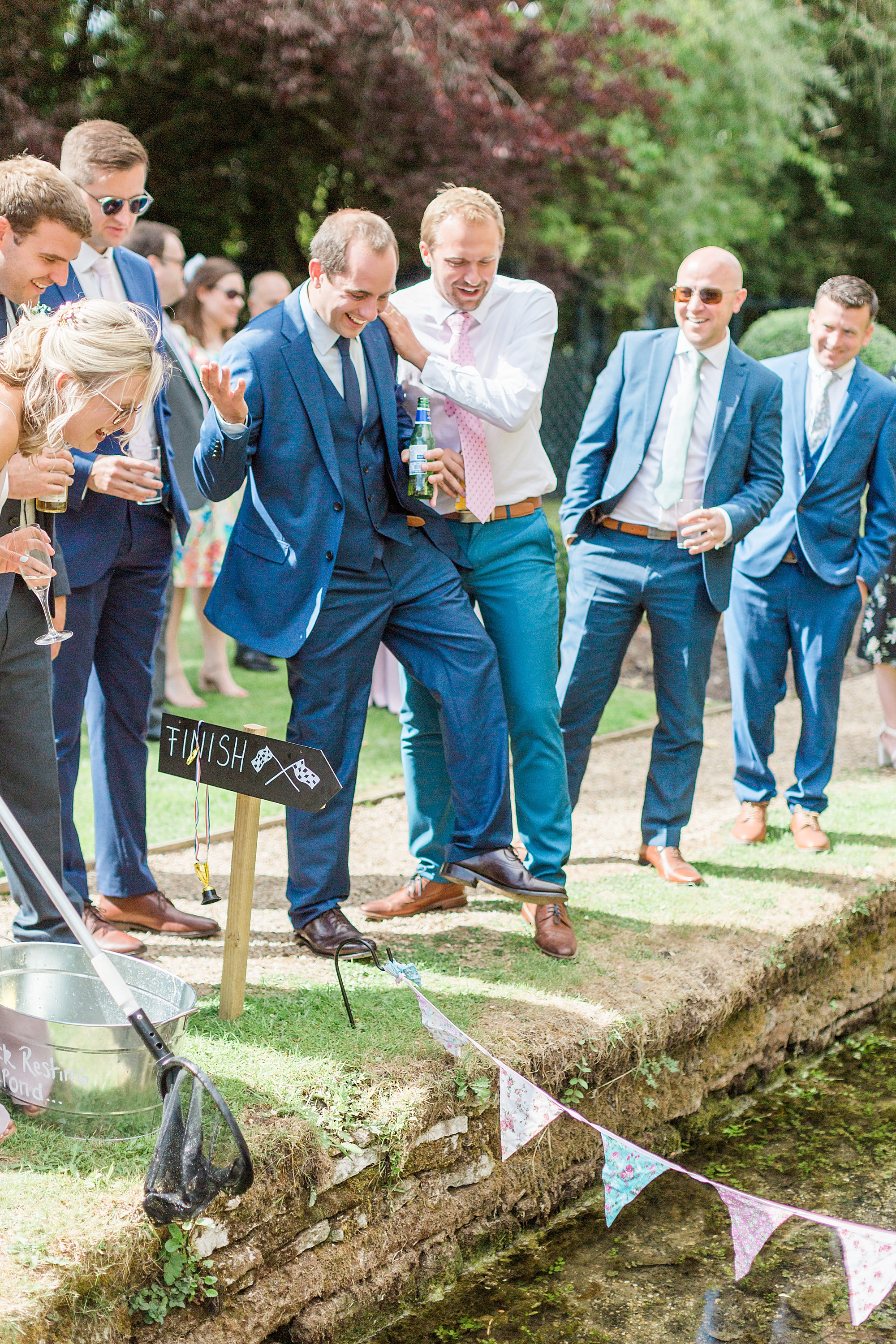image shows guests enjoying themselves at a wedding reception whilst racing rubber ducks down the stream at the swan at bibury in the cotswolds
