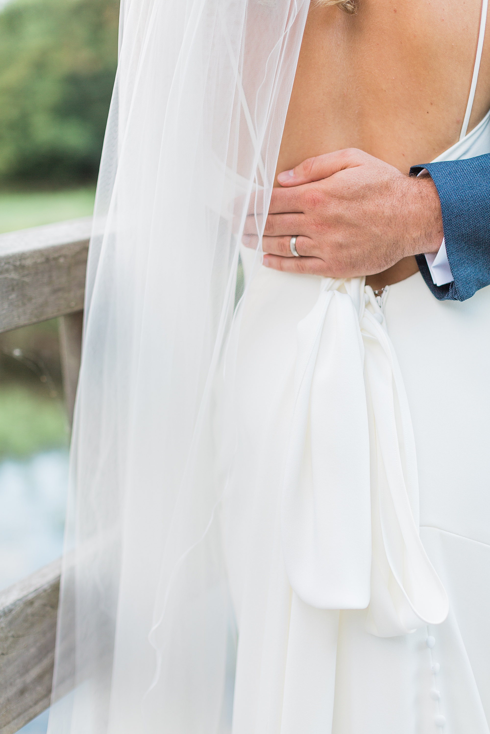photo shows a bride and groom stood on a bridge, the photo is close up of the bride's back and her dress and veil. the groom's left hand is around the bride's back and the photo focuses on his ring 