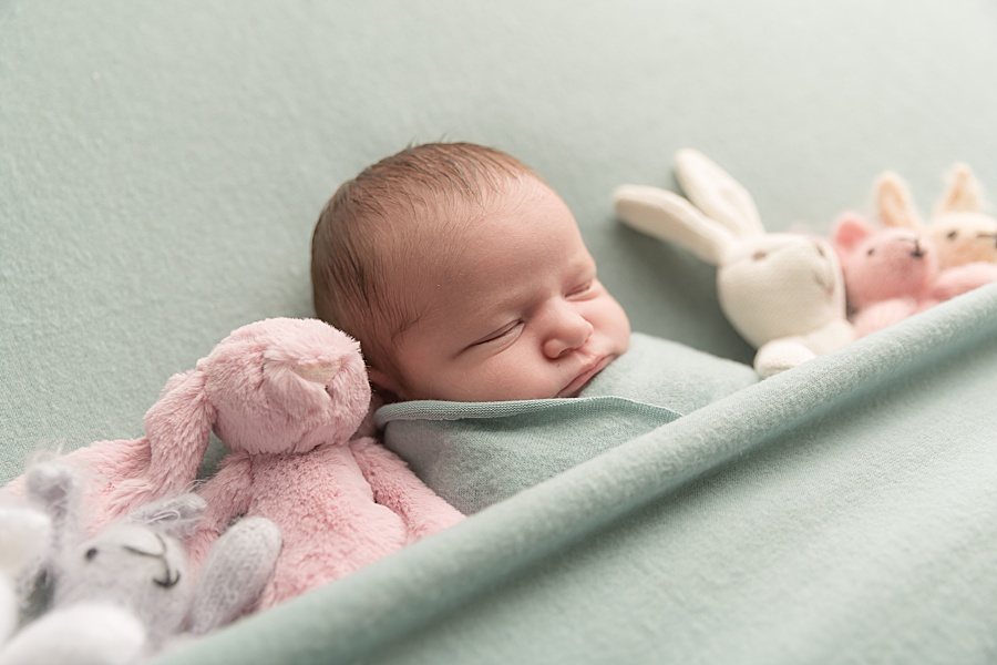 Photo by hayley morris Photography trained newborn photographer in malvern worcester. Image shows a newborn baby on a pale green fabric wrapped in a matching fabric. She's sleeping and has the fabric over her so that she looks like she's in a cot/bed sleeping. There are several pastel shade bunnies lay and tucked in either side of her 