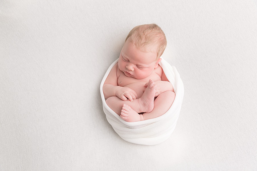 Image by hayley morris photography, trained newborn photographer specialist - image showing a newborn baby wrapped around her outer body and lay posed in a womb like position on a white fabric in a studio setting 
