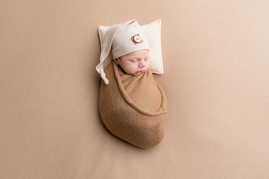 Image by hayley morris photography, trained newborn photographer specialist - image showing a newborn baby wrapped and lay on a neutral coffee coloured fabric with a sleepy cap and his head on a pillow