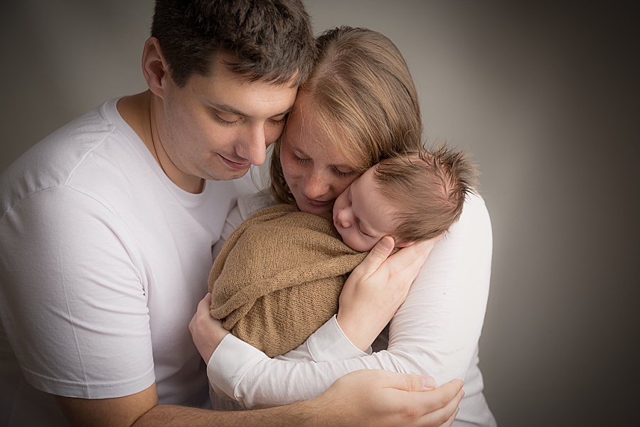 Image by hayley morris photography, trained newborn photographer specialist - image showing new family of mum, dad and baby boy wrapped, the three are traditionally posed against a neutral backdrop looking down towards their baby and cuddled in together 