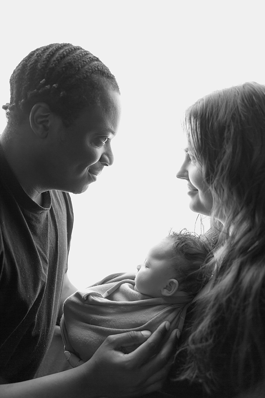 Image by hayley morris photography, trained newborn photographer specialist - image showing a newborn baby wrapped and held between their mum and dad during their newborn session. The image is black and white and the shot is backlit