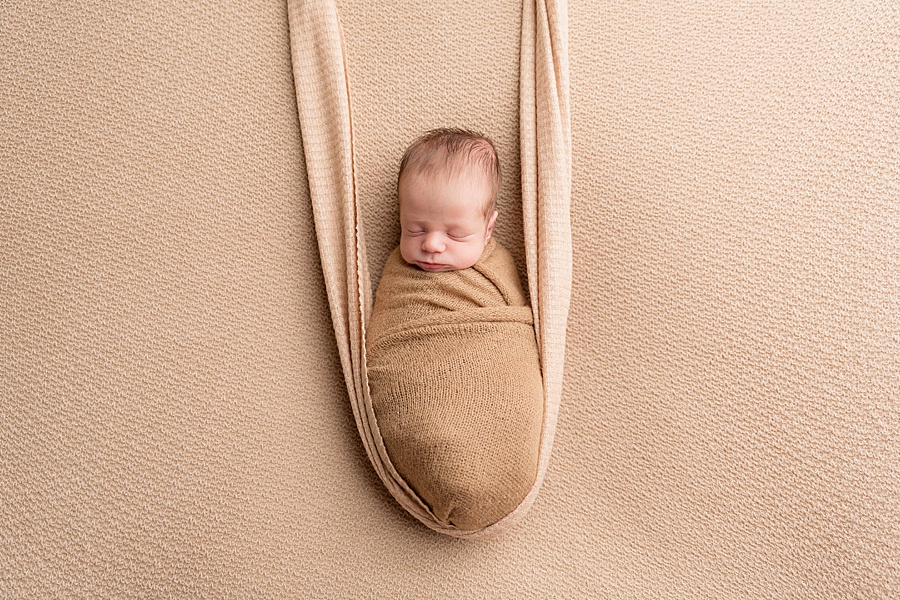 Image by hayley morris photography, trained newborn photographer specialist - image showing a newborn baby wrapped and posed on a latte coloured fabric looking like he is on a fabric swing/hammock