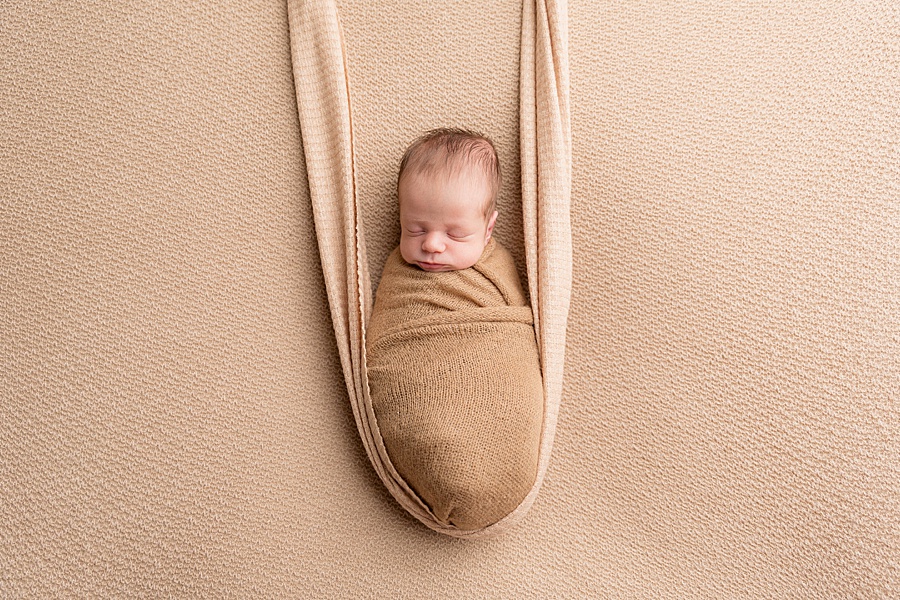Image by hayley morris photography, trained newborn photographer specialist - image showing a newborn baby wrapped and posed on a latte coloured fabric looking like he is on a fabric swing/hammock