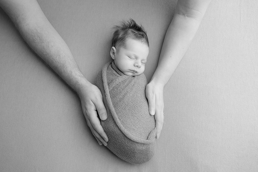 Photo captured by hayley morris photography trained newborn photographer in malvern worcestershire - image shows newborn baby wrapped in fabric and lay on a matching fabric with his parent's hands either side of him. Image is in black and and white 