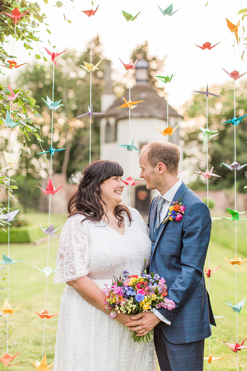 bride and groom stood in front of handmade origami cranes in the garden of homme house. The summerhouse is pictured in the background, evening sky 