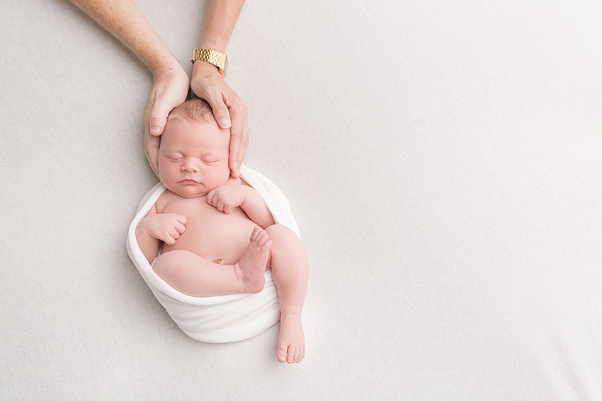 photo of a newborn baby curled up and loosely wrapped. she is posed on a creamy white backdrop and has each of her mums' hands on each side of her head 