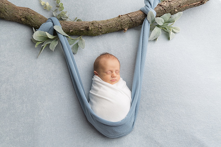 photo on a blue backdrop of a baby wrapped in a white wrap, posed in a blue fabric hammock and appears to be hanging from a branch with foliage. He's safely posed for newborn photography 