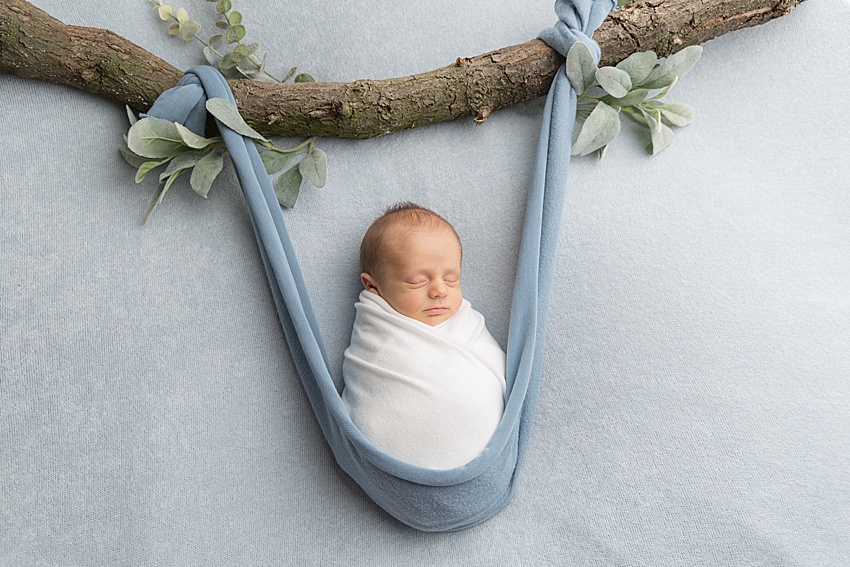 blue backdrop and a baby wrapped in a white wrap lay in a blue hammock. Baby appears to be posed with the hammock hanging from a large branch with foliage. All posing is done very safely for newborn photography 