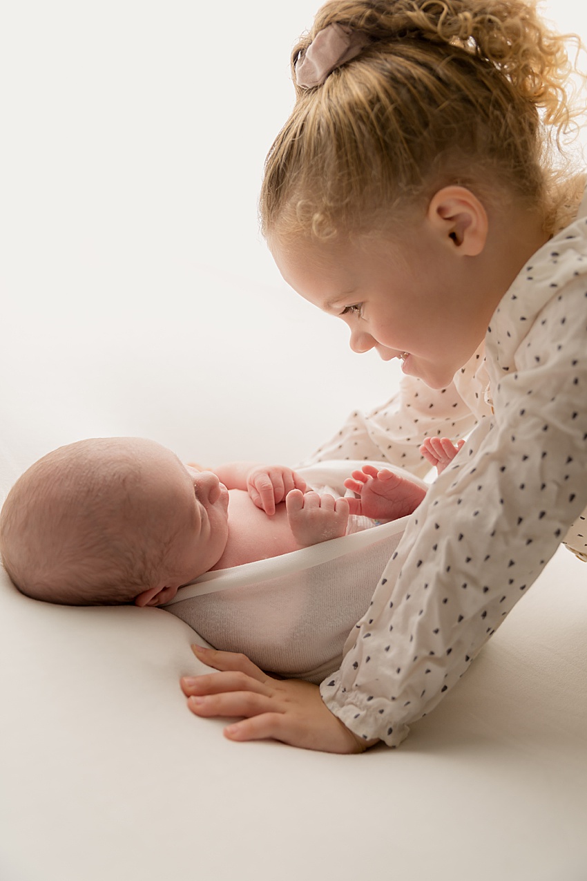 newborn baby lay loosely wrapped on a posing bag. baby is lay on his back and there is backlighting. Above him his older sister is looking down towards him smiling and very proud 