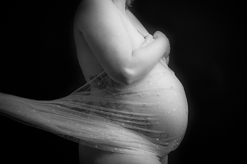 Black backdrop and a pregnancy lady photographed from the chest down with a sheer material covering her pregnant tummy. Image is in black and white 
