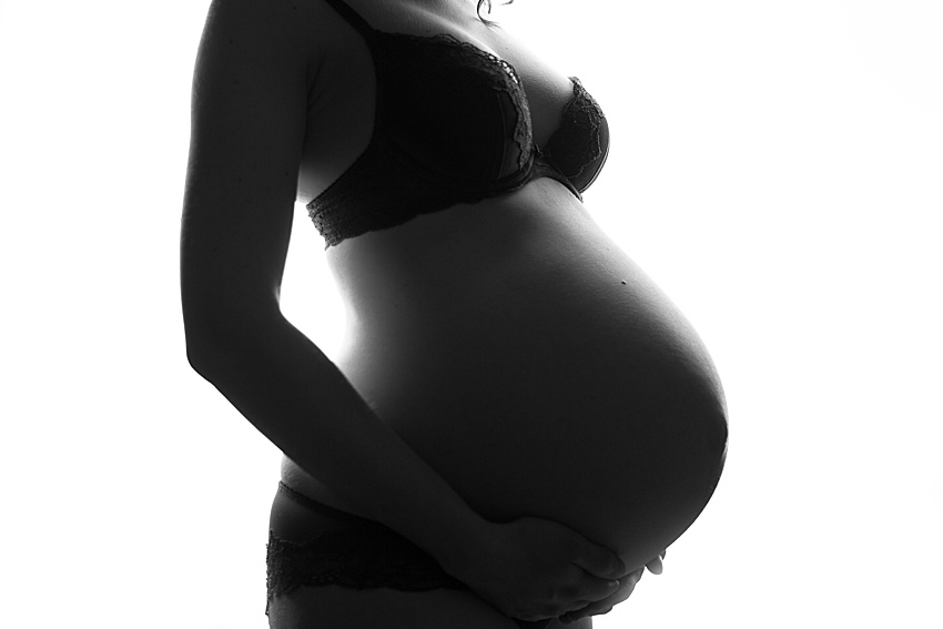 backlit silhouette photo in black and white of a woman's pregnant tummy photographed from her chest down to her lower bump. The lady is wearing underwear and her hand is under her bump 