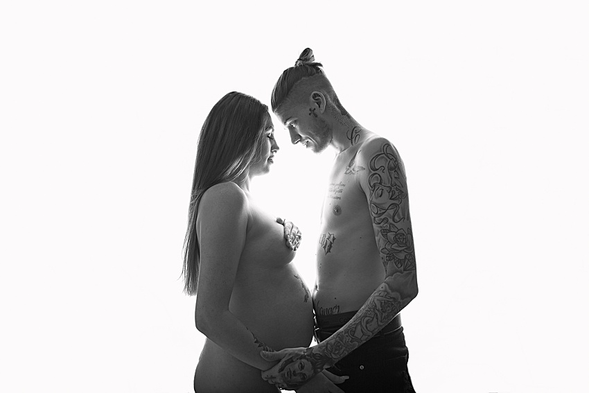 backlit photo of a couple facing in towards each other. the lady is pregnant and posed carefully nude, the man is posed shirtless. They are holding hands and the lady is posed with her arm over her chest