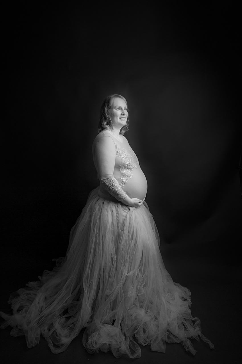 black and white photo of a pregnant lady wearing an embroidered bodysuit and tulle skirt, she's smiling and looking upwards 