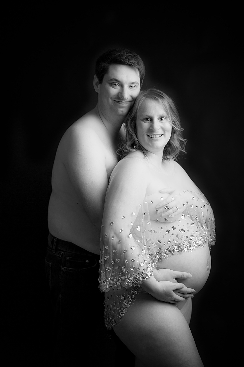 black and white photo of a couple posed together. the lady is expecting a baby wearing a sheer cape with jewels over it . her partner is stood behind her embracing her wearing jeans and shirtless 