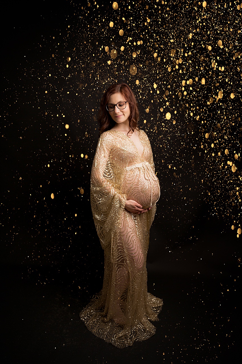 Pregnant lady photographed against a black backdrop wearing a gold glittering dress with long big draping sleeves. Lady is looking downwards to the side holding her pregnant tummy. The photo has added gold glitter in processing to add to the elegance and glamour of the image