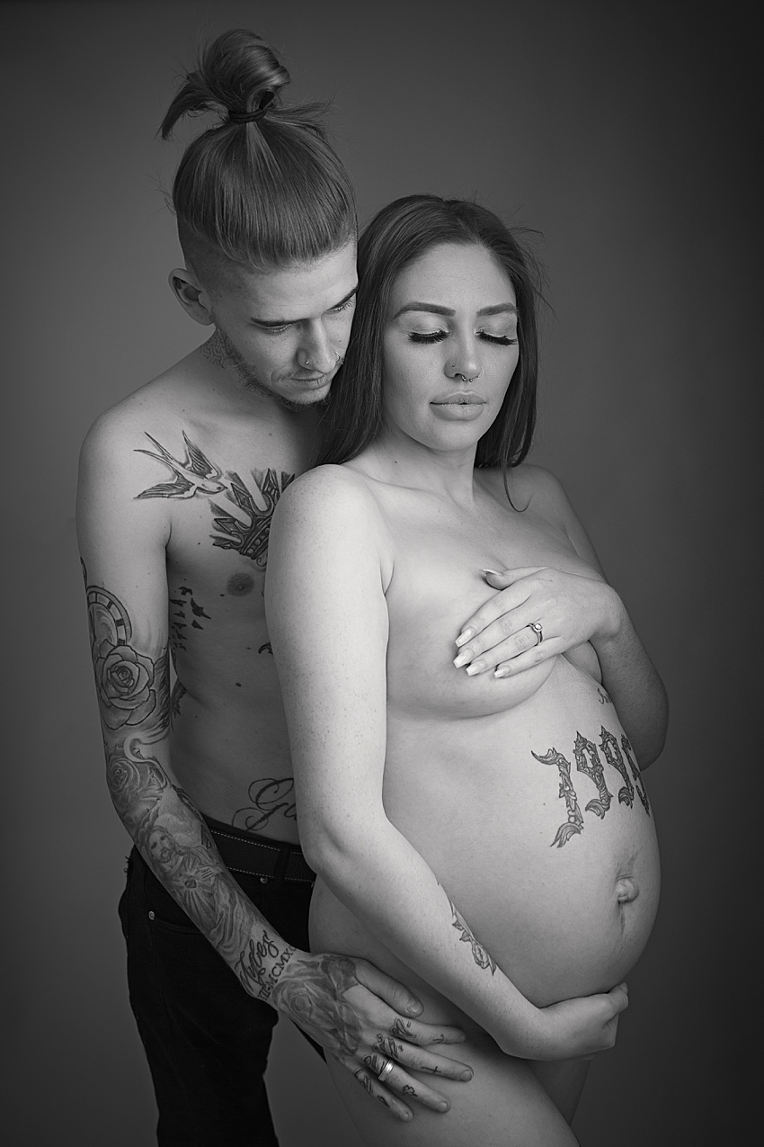 black and white photo of a man and woman posed together, the woman is nude, posed sensitively covering her chest and is expecting a baby. Her partner is stood embracing her with his hand on her hip looking down towards her 