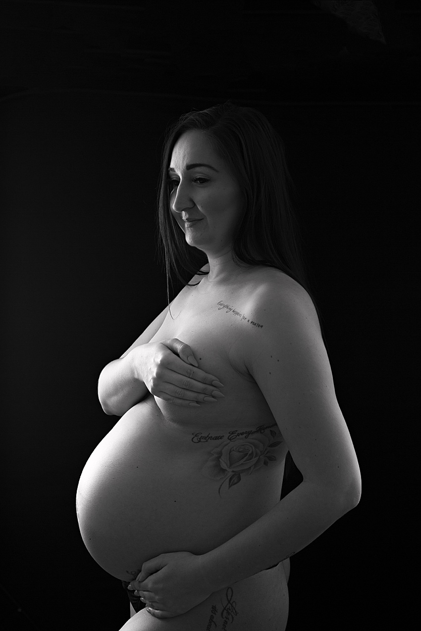 Photograph of a pregnant lady with tattoos posed nude sensitively with her hand over her chest. The light is very low and just skims over her pregnant tummy creating a lovely look 