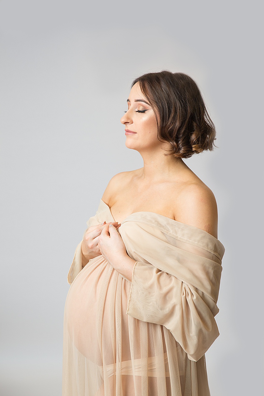 Pregnant lady wearing a nude colour sheer gown photographed for her maternity session against a grey backdrop