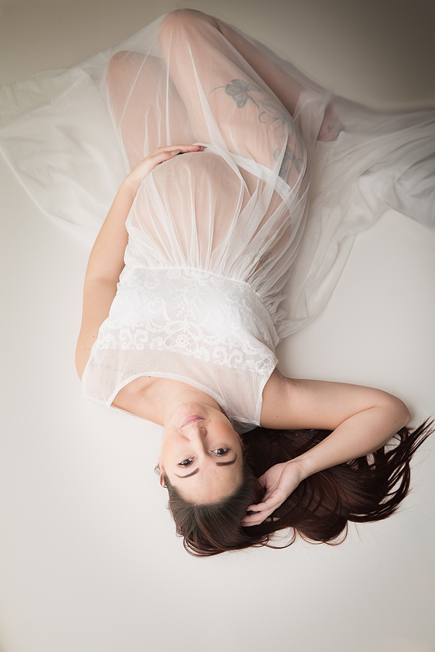 pregnant lady lay on the floor posed wearing a white dress with sheer lower half, she has her one hand under her tummy, knees bent inwards and one hand towards her head and her long brown hair posed around her 