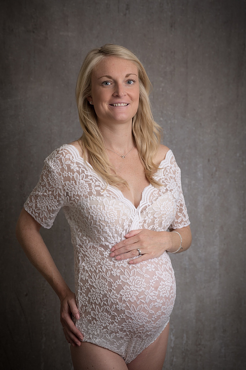 Photo of a pregnant lady posed wearing a white lace bodysuit holding her tummy, She's looking towards the camera smiling 