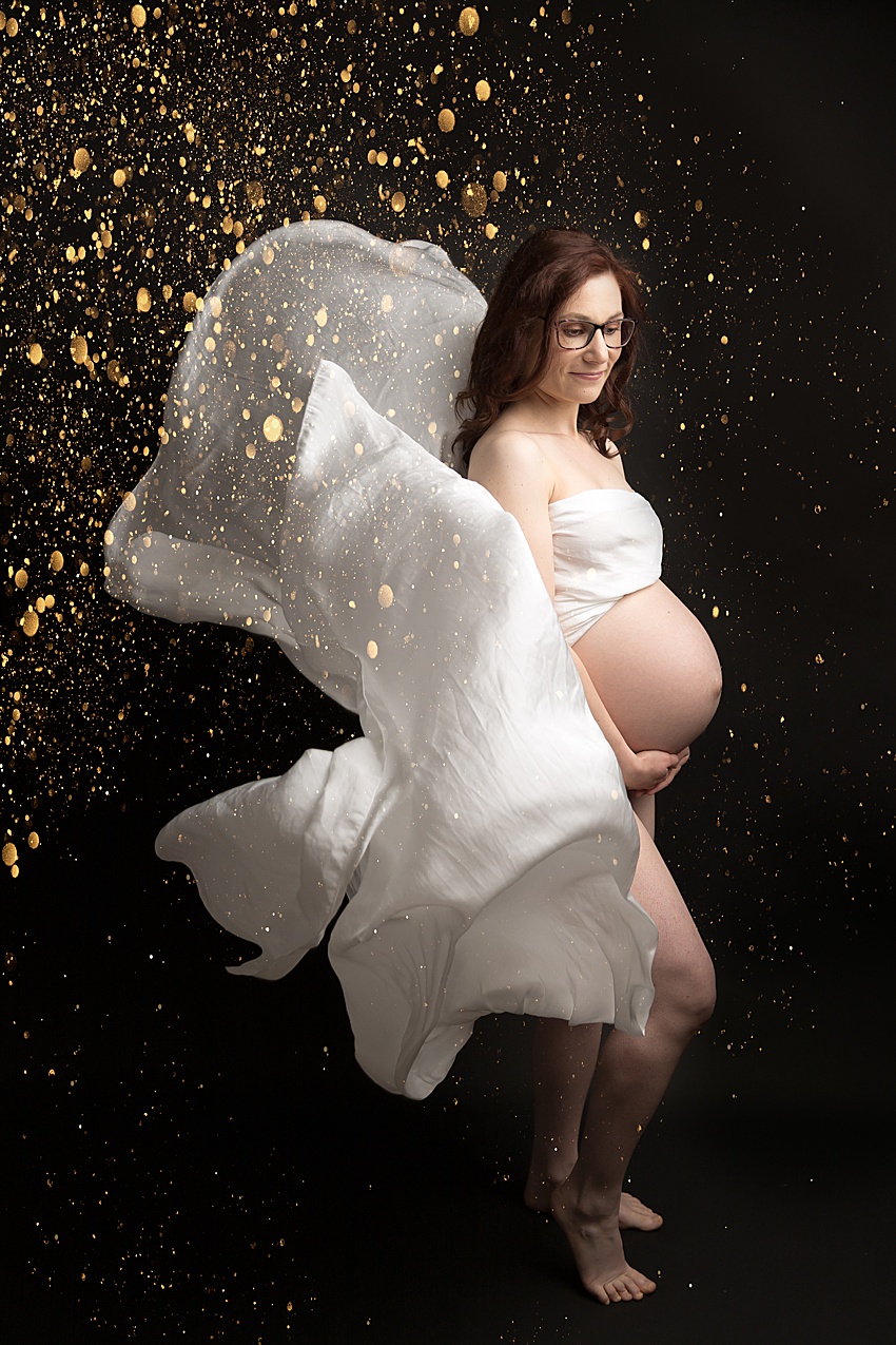 pregnant lady photographed against a black backdrop wrapped in a white silk scarf which has been thrown and floating down, gold glitter effect over the photo to one side  