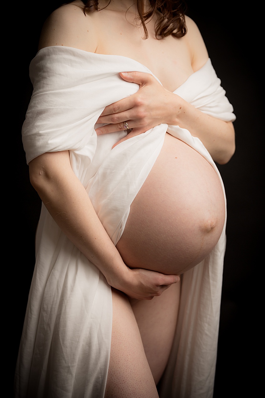 photograph of a pregnant lady photographed from the neck down holding silk material over her chest revealing her pregnant tummy.